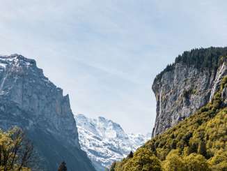 The Staubach Falls plunge over a high, steep rock face into the Lauterbrunnen Valley. In the foreground are green trees and meadows, while on the left, the view opens up between the rock faces to reveal the mountain landscape.