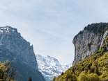 The Staubach Falls plunge over a high, steep rock face into the Lauterbrunnen Valley. In the foreground are green trees and meadows, while on the left, the view opens up between the rock faces to reveal the mountain landscape.