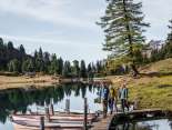 Drei Personen gehen über einen schmalen Holzsteg, an dem mehrere Ruderboote festgemacht sind. Der Engstlensee spiegelt die umliegenden Bäume und den Himmel klar wider. Die Umgebung wirkt herbstlich und ruhig.