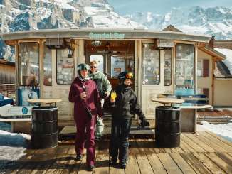 Drei Personen stehen vor der Gondelbar beim Hotel Jungfrau in Mürren und geniessen gemeinsam ein Getränk. Die sonnige Terrasse und die verschneite Bergkulisse schaffen eine gemütliche Après-Ski-Atmosphäre.