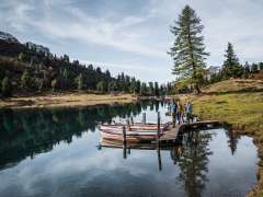 Trois personnes marchent sur une étroite passerelle en bois, à laquelle sont amarrées plusieurs barques à rames. Le lac d'Engstlen reflète clairement les arbres environnants et le ciel. Le cadre dégage une atmosphère automnale et paisible.
