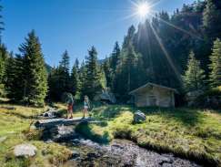 Zwei Wanderer unterwegs auf dem Säumerweg in Gadmen. Sie überqueren den Bergbach auf einer kleinen Holzbrücke vor einer Alphütte. Die Sonne scheint oberhalb der Tannen auf die Lichtung.