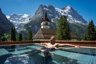A guest enjoys the outdoor pool at the Hotel Fiescherblick in Grindelwald. In the background, the Grindelwald village church and the striking Ostegg mountain form a stunning backdrop against a bright blue sky.