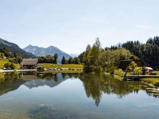 Der Blick über den Badesee zeigt grüne Wiesen, verstreute Häuser und Wälder im Hintergrund. In der Mitte spiegelt sich das kleine Holzhaus im Wasser, während die Berglandschaft die idyllische Szenerie vollendet.