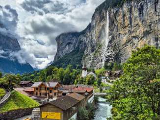 Vue sur le village de Lauterbrunnen, avec ses chalets et la Lütschine qui coule. À l'arrière-plan, la cascade du Staubbach se jette dans la vallée depuis la paroi rocheuse escarpée et marque le paysage du village.