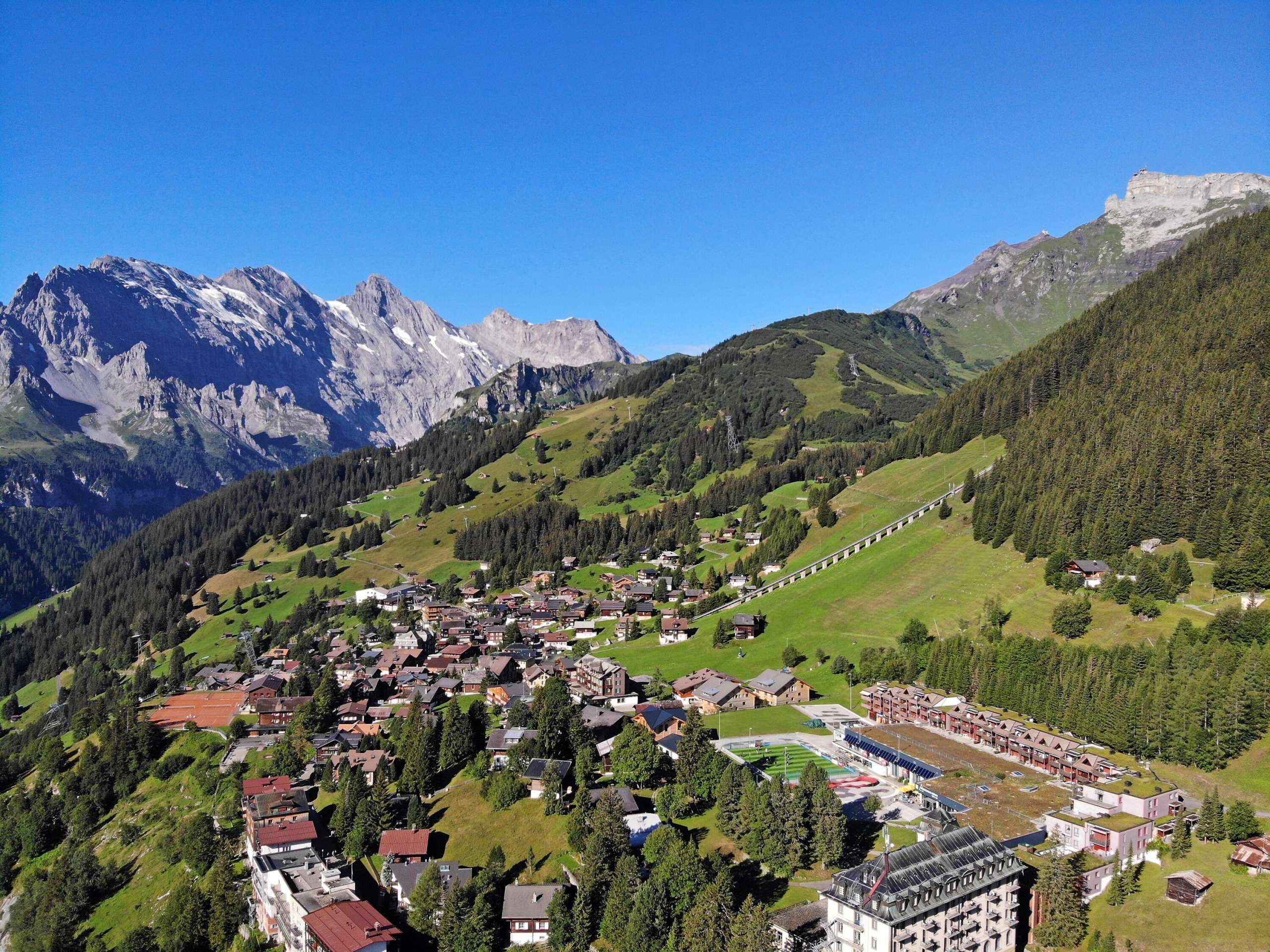 Mürren village Vue aérienne du village de Mürren. Le village est entouré de prairies et de forêts verdoyantes. En arrière-plan, on aperçoit le ciel bleu.