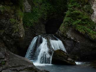 Ein Abschnitt des Reichenbachfalls stürzt in mehreren Strömen über dunkle Felsen in ein ruhiges Becken. Die enge Schlucht ist von moosbewachsenen Wänden, dichter Vegetation und überhängenden Ästen umgeben.