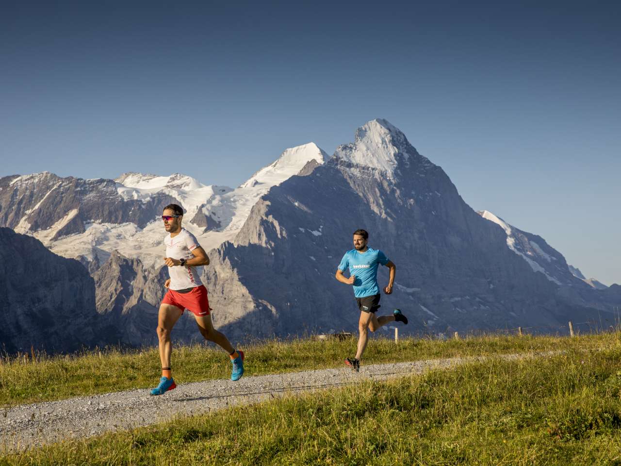 Deux personnes en train de faire du trail running sur un sentier de montagne, entourées d'un paysage alpin avec des rochers, des prairies et des montagnes en arrière-plan.