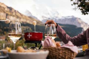 Ein rotes Fodue Caquelon steht auf einem Tisch im Außenbereich. Geschmolzener Käse wird mit Gabeln herausgehoben. Weingläser und Brotkorb stehen daneben. Im Hintergrund sind bewaldete Hänge und alpine Berge sichtbar.
