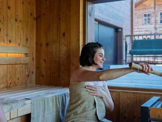 Two guests in the sauna at the Hotel Fiescherblick in Grindelwald, looking out through the panoramic window at the hotel terrace and the surrounding buildings.