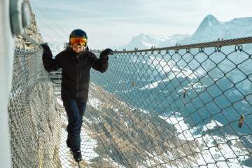 Eine Frau balanciert auf einem Drahtseil beim Felsensteg Thrill Walk mit eindrücklichem Winter-Bergpanorama mit dem Eiger im Hintergrund.