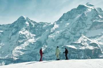Drei Wintersportler*innen stehen auf der Skipiste im Skigebiet Mürren–Schilthorn und geniessen den Ausblick auf Eiger, Mönch und Jungfrau. Die mächtige Bergkulisse und die verschneite Piste schaffen eine eindrucksvolle alpine Szenerie.