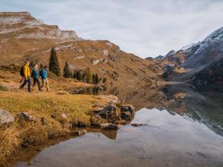 Drei Personen gehen am Ufer des Engstlensees entlang. Das Wasser ist ruhig und reflektiert die umliegenden Hänge und Felsformationen. Im Vordergrund liegen Steine und herbstliche Gräser.
