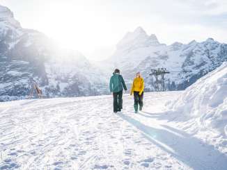 Two people are walking from the First mountain station toward Oberjoch along a snow-covered winter hiking trail. The low-hanging sun creates a bright backlight and long shadows in the snow. Striking, snow-capped mountain peaks are visible in the background.