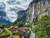 A view of the village of Lauterbrunnen with its chalets and the flowing Lütschine River. In the background, the Staubbach Falls plunge from the steep rock face into the valley, defining the village’s skyline.