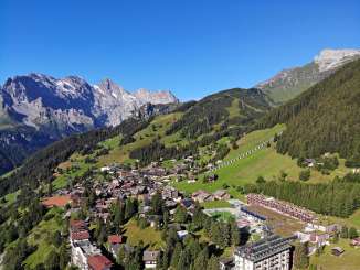 Luftaufnahme vom Dorf Mürren. Das Dorf ist umgeben von grünen Wiesen und Wälern. Im Hintergrund ist der blaue Himmel zu sehen.