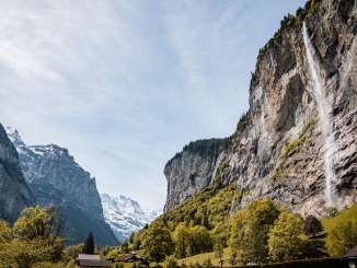 Der Staubachfall stürzt über eine hohe, steile Felswand ins Lauterbrunnental. Im Vordergrund stehen grüne Bäume und Wiesen, während sich links der Blick zwischen den Felswänden in die Berglandschaft öffnet.