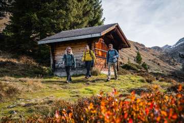 Three hikers walk past a small wooden cabin standing in an open, gently sloping meadow. In the background, bare mountain slopes and the first patches of snow are visible. The scene depicts a peaceful, natural mountain setting.