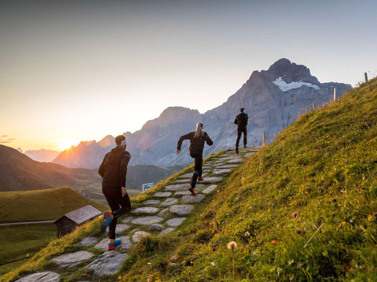 Trois personnes en train de faire du trail running sur un sentier de montagne, entourées d'un paysage alpin avec des rochers, des prairies et des montagnes en arrière-plan.