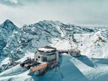 Die Drohnenaufnahme zeigt das Schilthorn mit dem markanten Drehrestaurant Piz Gloria und der Bergstation, eingebettet in die verschneite Hochgebirgslandschaft. Rund um den Gipfel erstrecken sich die schneebedeckten Bergketten der Berner Alpen und verleihen der Szenerie ihre eindrucksvolle Weite.