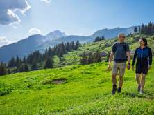 Deux randonneurs marchent sur un sentier à travers les prairies de la Grande Scheidegg. Entourés de prairies alpines d'un vert luxuriant et de forêts de montagne denses, ils profitent de la vue sur le paysage montagneux environnant sous un ciel ensoleillé parsemé de quelques nuages épars.