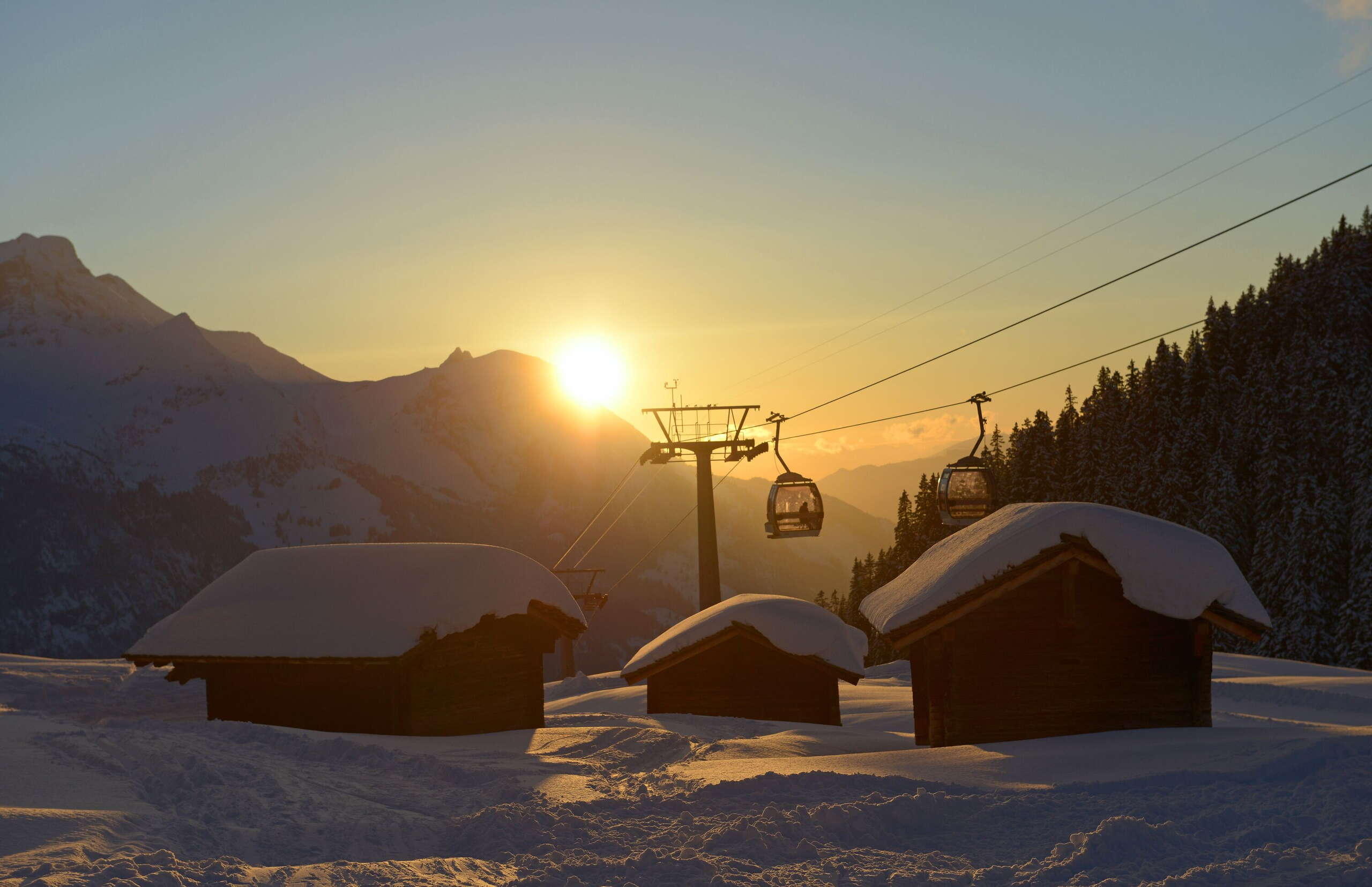 Zwei Gondeln der Bergbahnen Meiringen-Hasliberg im Sonnenuntergang.Im Vordergrund drei schneebedeckte Hütten. Aufgenommen bei der Zwischenstation Bidmi.