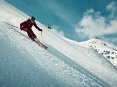 Skifahrerin bei einem sportlichen Schwung auf einer verschneiten Piste mit aufwirbelndem Schnee und Bergkulisse.