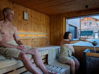 Two guests in the sauna at the Hotel Fiescherblick in Grindelwald, looking out through the panoramic window at the hotel terrace and the surrounding buildings.