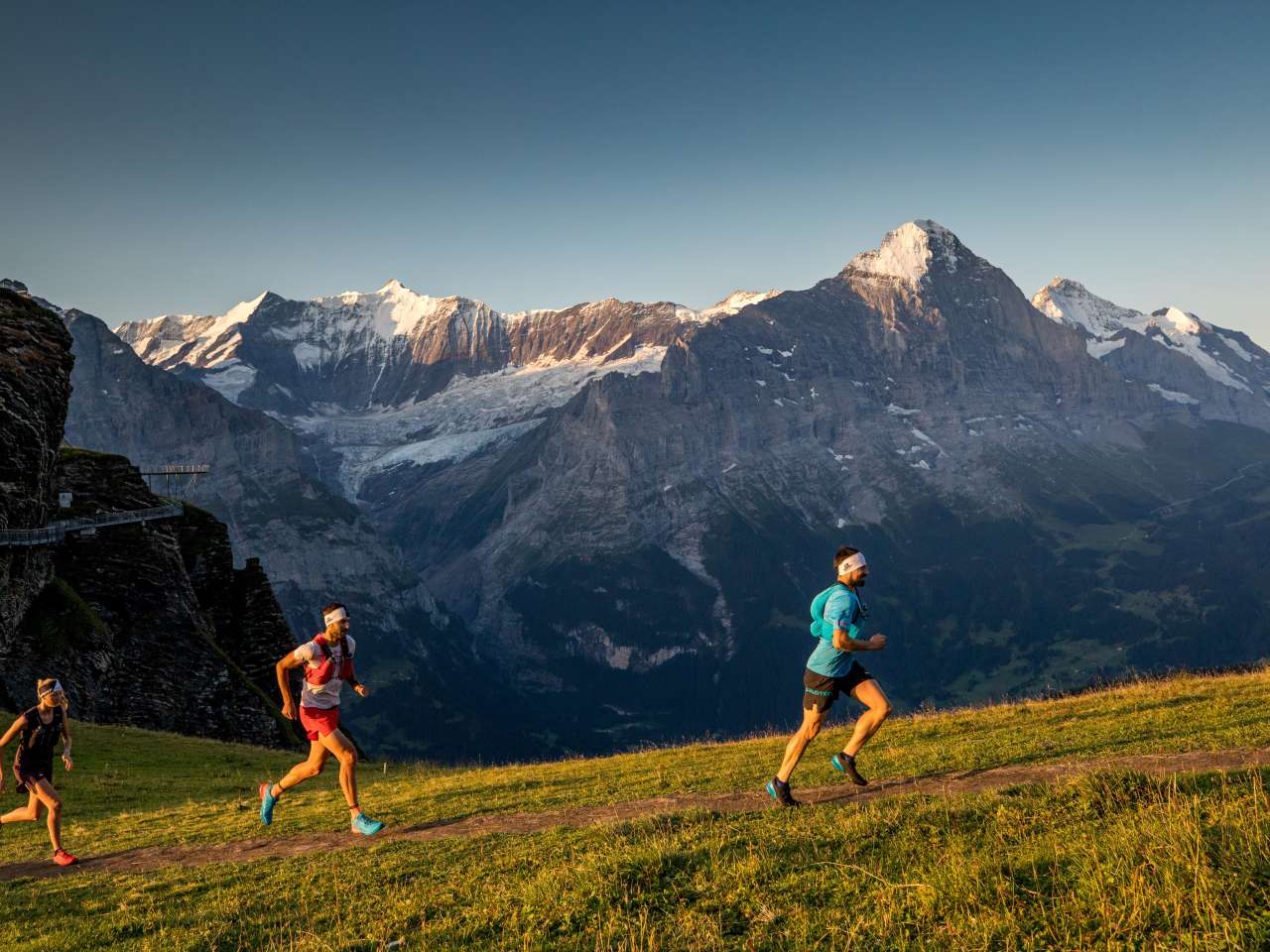 Trois personnes en train de faire du trail running sur un sentier de montagne, entourées d'un paysage alpin avec des rochers, des prairies et des montagnes en arrière-plan.