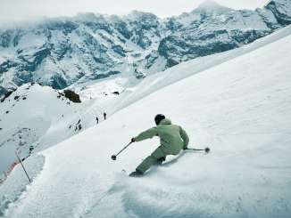 Skifahrer bei einem dynamischen Carving-Schwung auf einer verschneiten Piste mit aufwirbelndem Schnee und Bergkulisse.