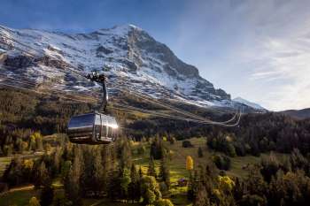 Le téléphérique de l'Eiger Express au printemps, avec un paysage verdoyant au premier plan, des montagnes enneigées en arrière-plan et un ciel bleu.