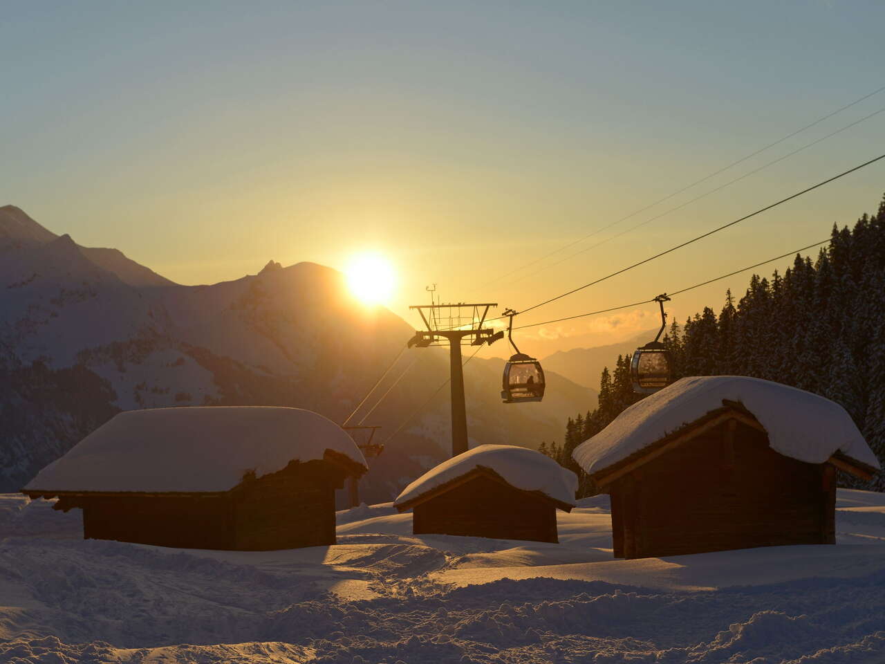 Deux télécabines des remontées mécaniques de Meiringen-Hasliberg au coucher du soleil, avec au premier plan trois cabanes enneigées. Prise de vue à la station intermédiaire de Bidmi.