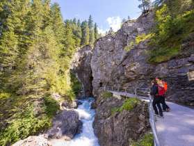 Au fil des siècles, la rivière a sculpté les gorges du glacier de Rosenlaui. Le sentier longe la paroi rocheuse qui surplombe la rivière et s'enfonce dans les gorges. Deux randonneurs admirent la rivière à l'entrée des gorges.