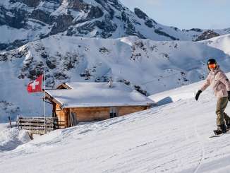 A snowboarder glides down the slope, while in the background a cozy mountain cabin flying the Swiss flag and the impressive Alpine scenery can be seen.