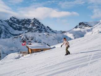 Une snowboardeuse dévale la piste, tandis qu'à l'arrière-plan on aperçoit un chalet de montagne accueillant, orné d'un drapeau suisse, et le magnifique panorama alpin.