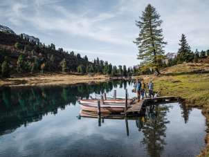 Drei Personen gehen über einen schmalen Holzsteg, an dem mehrere Ruderboote festgemacht sind. Der Engstlensee spiegelt die umliegenden Bäume und den Himmel klar wider. Die Umgebung wirkt herbstlich und ruhig.