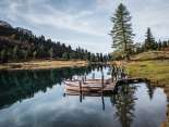 Drei Personen gehen über einen schmalen Holzsteg, an dem mehrere Ruderboote festgemacht sind. Der Engstlensee spiegelt die umliegenden Bäume und den Himmel klar wider. Die Umgebung wirkt herbstlich und ruhig.