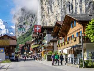 The village of Lauterbrunnen is surrounded by imposing cliffs. Tourists stroll through the village. The Staubbach Falls can be seen in the background.