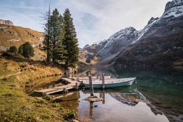 Mehrere Ruderboote liegen an einem schmalen Holzsteg am klaren Wasser des Engstlensees. Hohe Nadelbäume und herbstliche Hänge rahmen die Uferlandschaft. Im Hintergrund erheben sich steile, teils verschneite Berghänge.