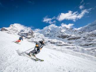 Two skiers are skiing down a groomed slope. A snow-covered mountain backdrop can be seen in the background.