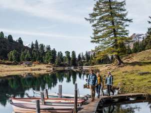 Trois personnes marchent sur une étroite passerelle en bois, à laquelle sont amarrées plusieurs barques à rames. Le lac d'Engstlen reflète clairement les arbres environnants et le ciel. Le cadre dégage une atmosphère automnale et paisible.