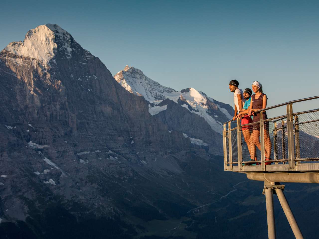 Trois personnes se tiennent sur une plate-forme d'observation, avec en arrière-plan un impressionnant décor de montagnes sous un ciel dégagé.