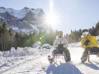 Zwei Personen schlitteln Richtung Schwarzwaldalp, während die tief stehende Sonne lange Schatten über den Schnee wirft. Im Hintergrund sind Bäume und das schneebedeckte Wetterhorn sichtbar.