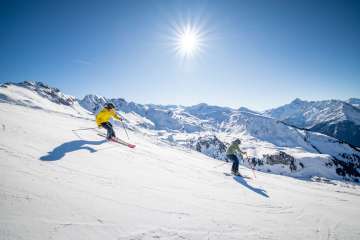 Zwei Skifahrer fahren über eine breite, verschneite Piste auf dem Hasliberg. Die Sonne steht hoch am Himmel und wirft lange Schatten auf den Schnee. Im Hintergrund sind weitere Bergketten und verschneite Gipfel sichtbar.