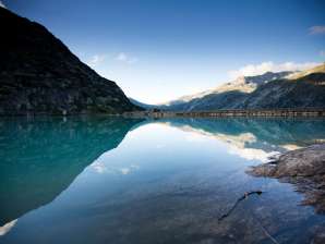 Le lac de Gelmer est niché au cœur du paysage de haute montagne du Grimsel. La surface calme de l'eau reflète les flancs des montagnes environnantes et le ciel limpide. Au premier plan, on aperçoit les rochers des rives, tandis qu'au fond, le barrage délimite le lac.