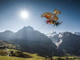 Der First Glider fliegt über eine grüne Wiese, im Hintergrund erheben sich Berge unter einem klaren blauen Himmel.