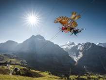 Der First Glider fliegt über eine grüne Wiese, im Hintergrund erheben sich Berge unter einem klaren blauen Himmel.
