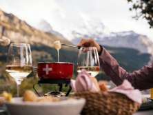 Ein rotes Fodue Caquelon steht auf einem Tisch im Außenbereich. Geschmolzener Käse wird mit Gabeln herausgehoben. Weingläser und Brotkorb stehen daneben. Im Hintergrund sind bewaldete Hänge und alpine Berge sichtbar.