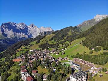 Vue aérienne du village de Mürren. Le village est entouré de prairies verdoyantes et de forêts. On aperçoit le ciel bleu en arrière-plan.