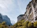 Der Staubachfall stürzt über eine hohe, steile Felswand ins Lauterbrunnental. Im Vordergrund stehen grüne Bäume und Wiesen, während sich links der Blick zwischen den Felswänden in die Berglandschaft öffnet.
